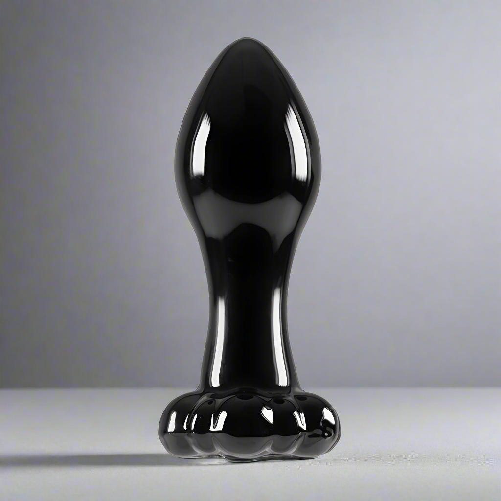 The NS Novelties Crystal Flower Butt Plug is a shiny black, cone-shaped borosilicate glass object that stands upright on flat surfaces. Its bulbous top and flared base resemble a polished sculpture, while its versatile shape complements any soft gray backdrop.