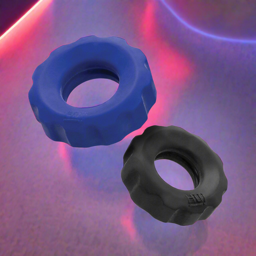 Two circular Hunky Junk Cog Rings, one blue and one black, are displayed on a reflective surface with pink and purple background lights. Made from plus+silicone TPR/silicone blend, these c-rings in the Hunky Junk Cog Ring 2 Size Double Pack feature ridged exteriors and hollow centers.