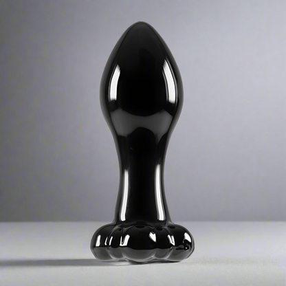 The NS Novelties Crystal Flower Butt Plug is a shiny black, cone-shaped borosilicate glass object that stands upright on flat surfaces. Its bulbous top and flared base resemble a polished sculpture, while its versatile shape complements any soft gray backdrop.