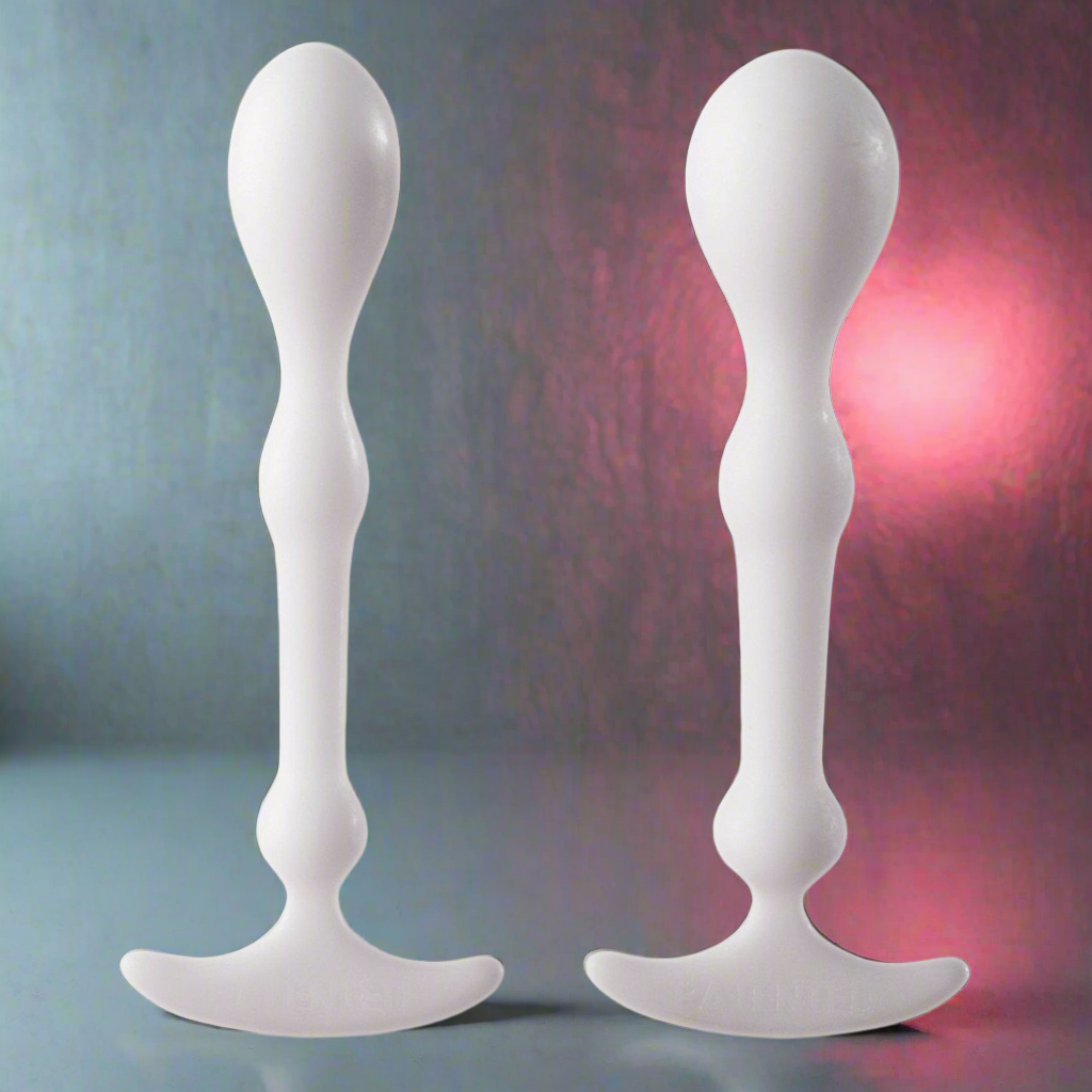 Two white, ergonomically shaped plastic devices from the Aneros Peridise Set stand upright on a reflective gray surface. They feature bulbous ends with a curved base. A soft pink light in the background creates an intimate ambiance for anal play.