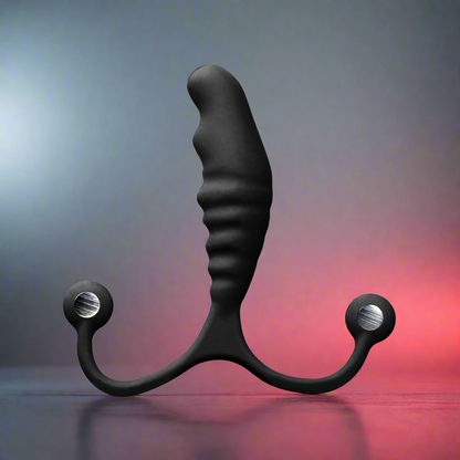 The Aneros Psy Adjustable Prostate Stimulator, featuring a black anatomical design with a wavy textured handle and two rounded arms with silver accents, is upright on a reflective surface. The background transitions from dark to light shades with subtle red highlights.