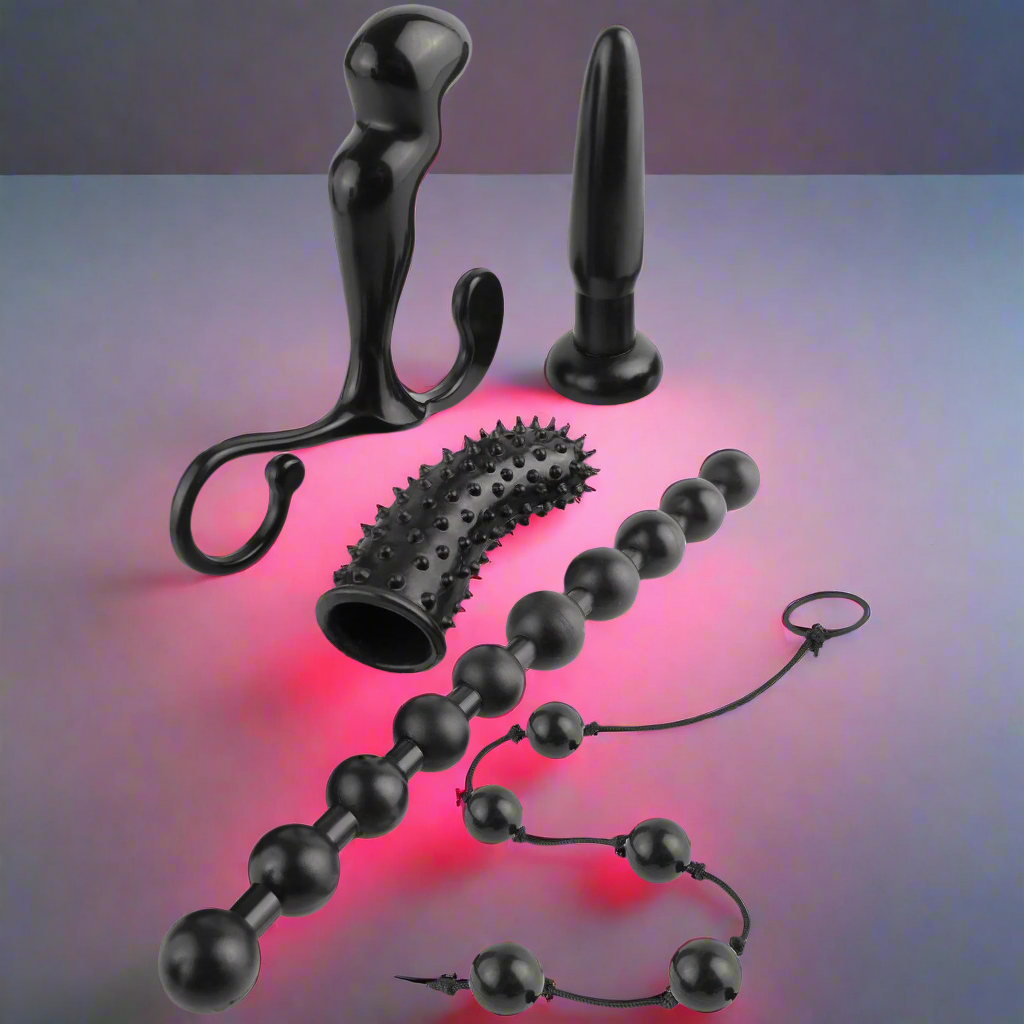 The Anal Fantasy Collection Beginners Fantasy Kit by Pipedream features black silicone adult toys arranged on a gradient surface with a red glow, showcasing various shapes and sizes like beads, a spiky sleeve, and plug-like items for diverse anal pleasures.