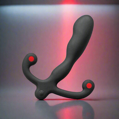 The Aneros Helix Syn V Prostate Massager, a black precision tool with a curved design and two small red accents on the handle, rests on a reflective surface. It is softly illuminated by red light against a gradient gray background.