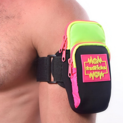 A person wears the Ekg Arm Bag in Neon by *ssTricks in green, pink, and black, featuring reflective side paneling, two zippered pockets, and a “*ssTricks” patch on the upper arm. The background is plain white.