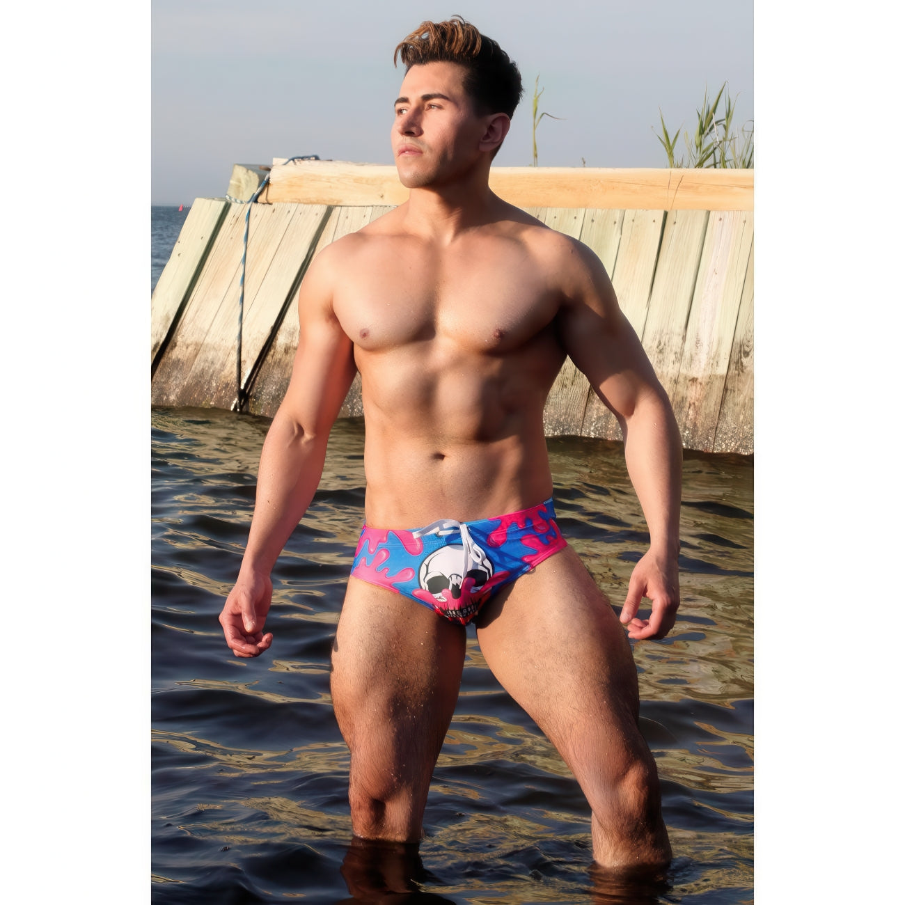A muscular man stands in shallow water by a wooden dock, wearing the *ssTricks Fantasy Swim Brief - Skull Pop. He looks confidently to the side, sunlight highlighting his upper body and radiating effortless beach style.