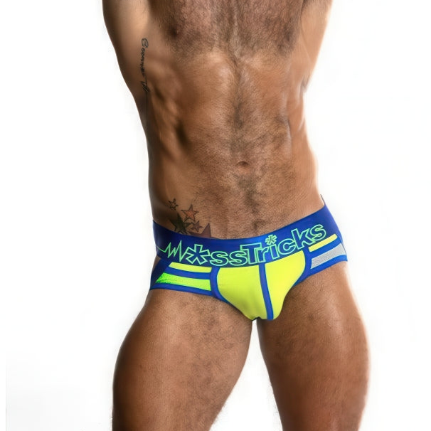 A shirtless person models vibrant yellow and blue JUKU JOCK ExtraTerrestrial underwear by *ssTricks, featuring the brand name on the waistband. Tattoos adorn their left torso against a plain white background, highlighting their upper legs and torso.