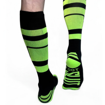 A person sports *ssTricks KNEE HIGH SOCKS in black and neon green with bold stripes. The back view highlights a lifted foot showing the soles stylized text design, adding edgy flair against a plain white background. These vibrant socks even feature hidden pockets for extra functionality.