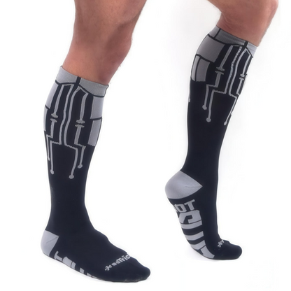 A person wearing *ssTricks CyberTech Socks & Black in knee-high length with gray and white geometric patterns, featuring branded text on the toes and soles, shown from side and front views on a white background.