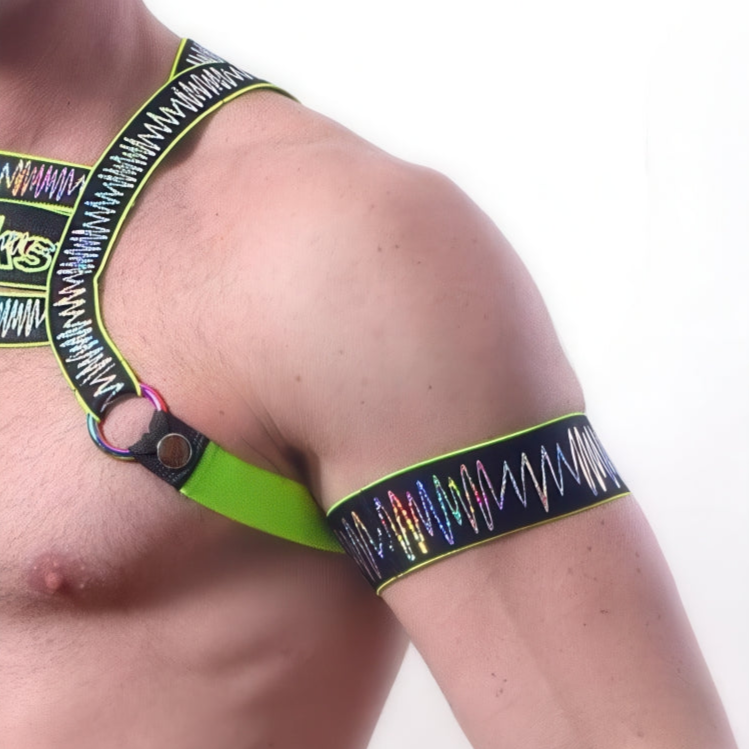 A person models the *ssTricks Electro Arm Bands in Neon Green, paired with a vibrant zigzag-patterned harness featuring metallic rings, highlighting the accessories on their shoulder and chest.