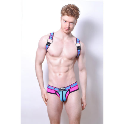 A muscular man with fair skin and curly red hair poses against a white background wearing *ssTricks Comicaze Brief Pink & Blue: vibrant, form-fitting neon pink mesh briefs with blue accents.