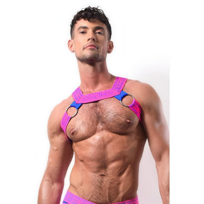 A muscular man with short dark hair confidently models the *ssTricks CyberTech Harness in Neon Pink & Blue, featuring metallic rings and a futuristic tech vibe, over his bare chest against a plain white background.
