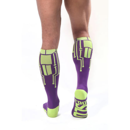 A person, back to the camera, wears *ssTricks CyberTech Socks Neon Yellow & Purple—purple knee-highs with neon yellow circuit designs and glowing heels/toes. FOOT is visible on one sock sole.