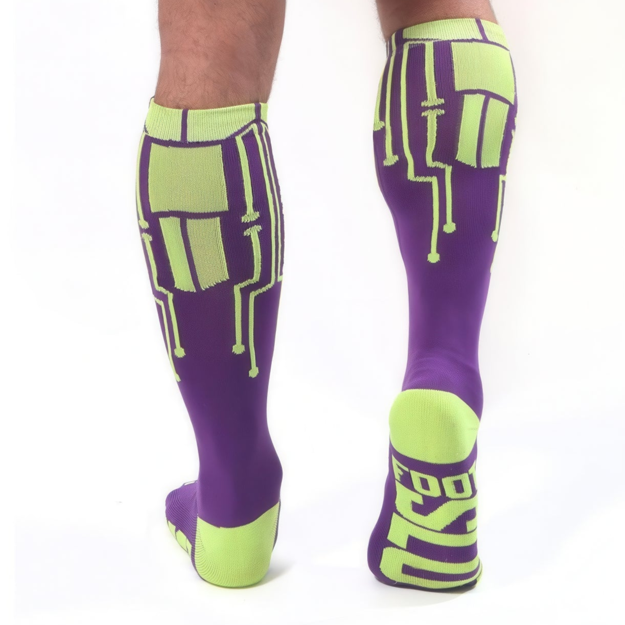 A person wears knee-high *ssTricks CyberTech Socks Neon Yellow & Purple with circuit board designs; one sole reads FOOT in bold lettering. A white background highlights their futuristic, tech-inspired style.