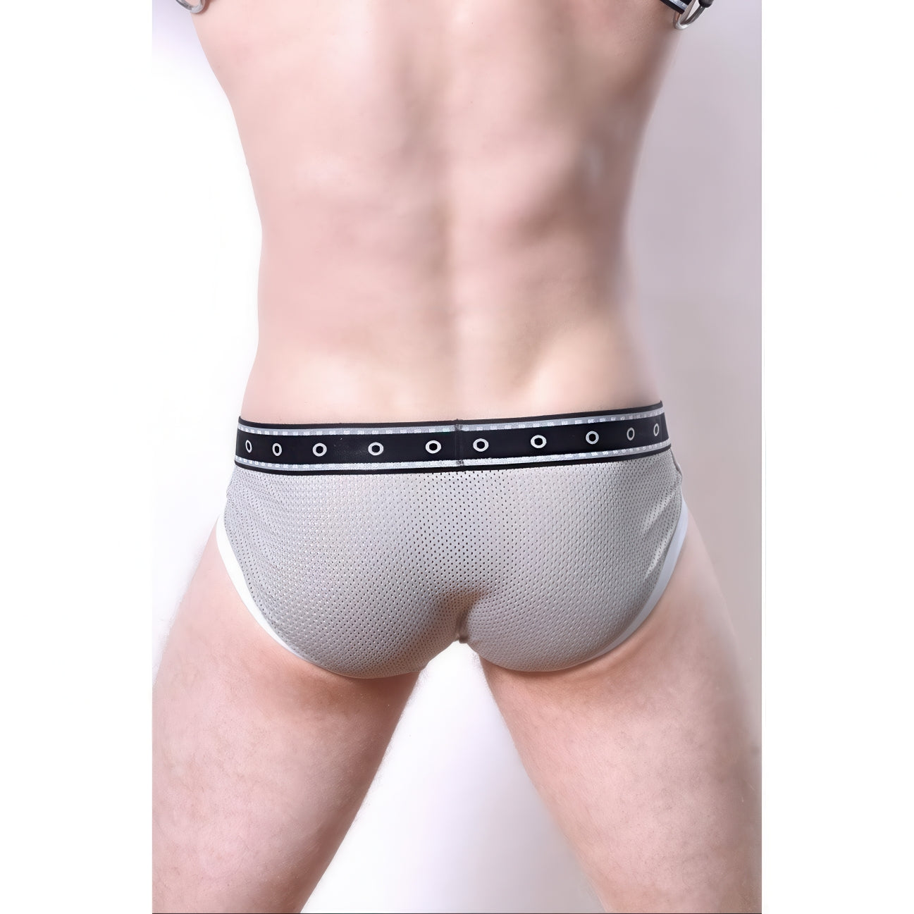 A person faces away from the camera, wearing *ssTricks Comicaze Brief Black & Grey mesh underwear with a black waistband and metal eyelets. The plain, light background highlights this blend of comfort and style.