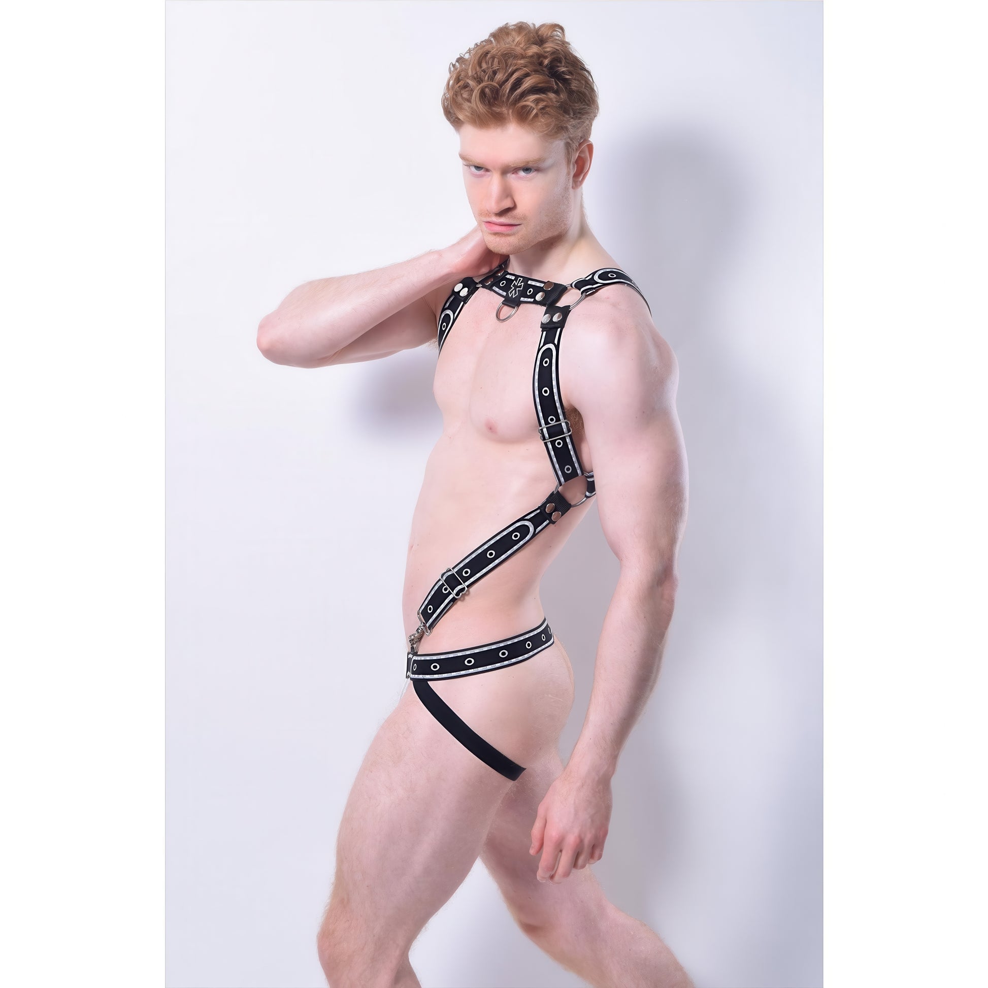A man with fair skin and curly red hair poses in an *ssTricks Comicaze Harness Black & Grey with detachable straps and a black strappy jockstrap, set against a plain light background.