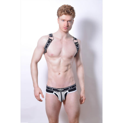 A muscular, light-skinned man with red hair poses against a white background, wearing an *ssTricks Comicaze Harness Black & Grey with detachable straps and matching underwear.