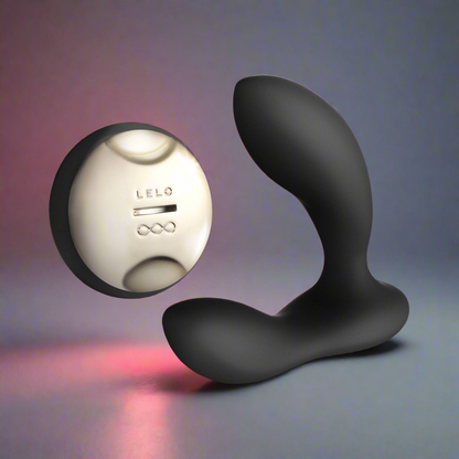 The LELO HUGO, a sleek black curved prostate massager, stands upright next to a round silver wireless remote labeled LELO. Both are placed on a smooth surface under soft gradient lighting.