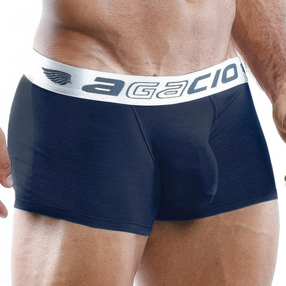 A close-up image shows a person wearing Agacio Boxer Trunks in black, featuring a white waistband displaying the brand name. The silky fabric and contouring pouch subtly accentuate their muscular build, capturing an elegant presentation of the midsection.