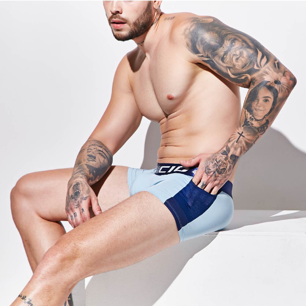 A tattooed man, shirtless, sits on a white surface against a white background. He wears supportive and comfortable Agacio Boxer Trunks in light blue and navy from the Agacio brand, with his right arm resting on his knee as he looks to the side.
