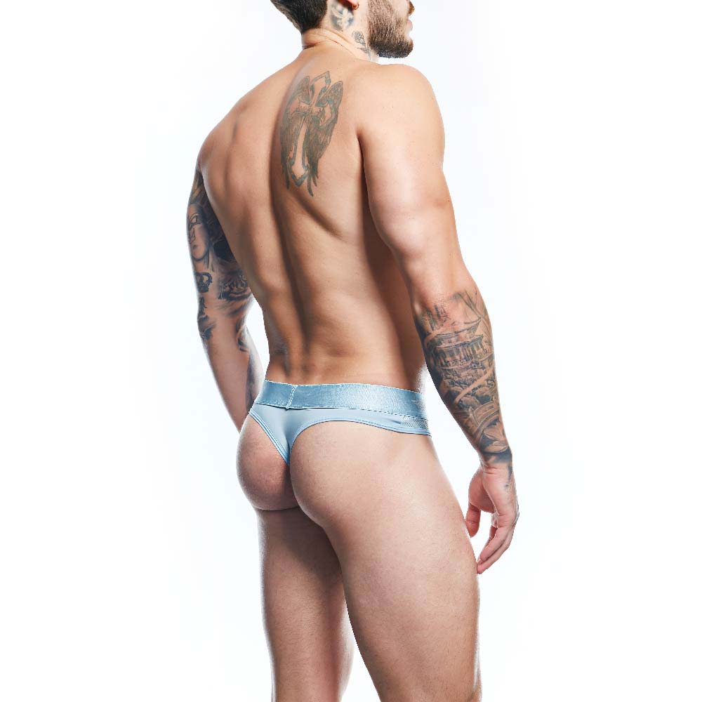 A shirtless, tattooed man stands sideways in Agacio Thong for Men, showcasing its minimalist design and premium fabric blend that highlights his toned physique against a white backdrop.