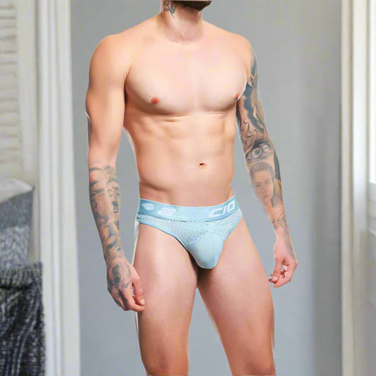 A muscular individual confidently poses in Agacio Thong for Men, made from premium fabric. Their arm tattoos stand out in a room with neutral tones and blurred furniture, highlighting the sleek design of their Agacio attire.