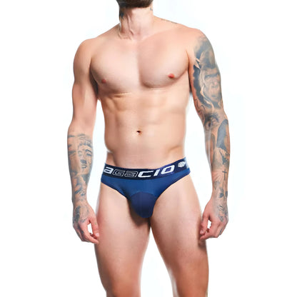 A tattooed man, shirtless and relaxed, faces forward wearing the Agacio Thong for Men on a plain white background.