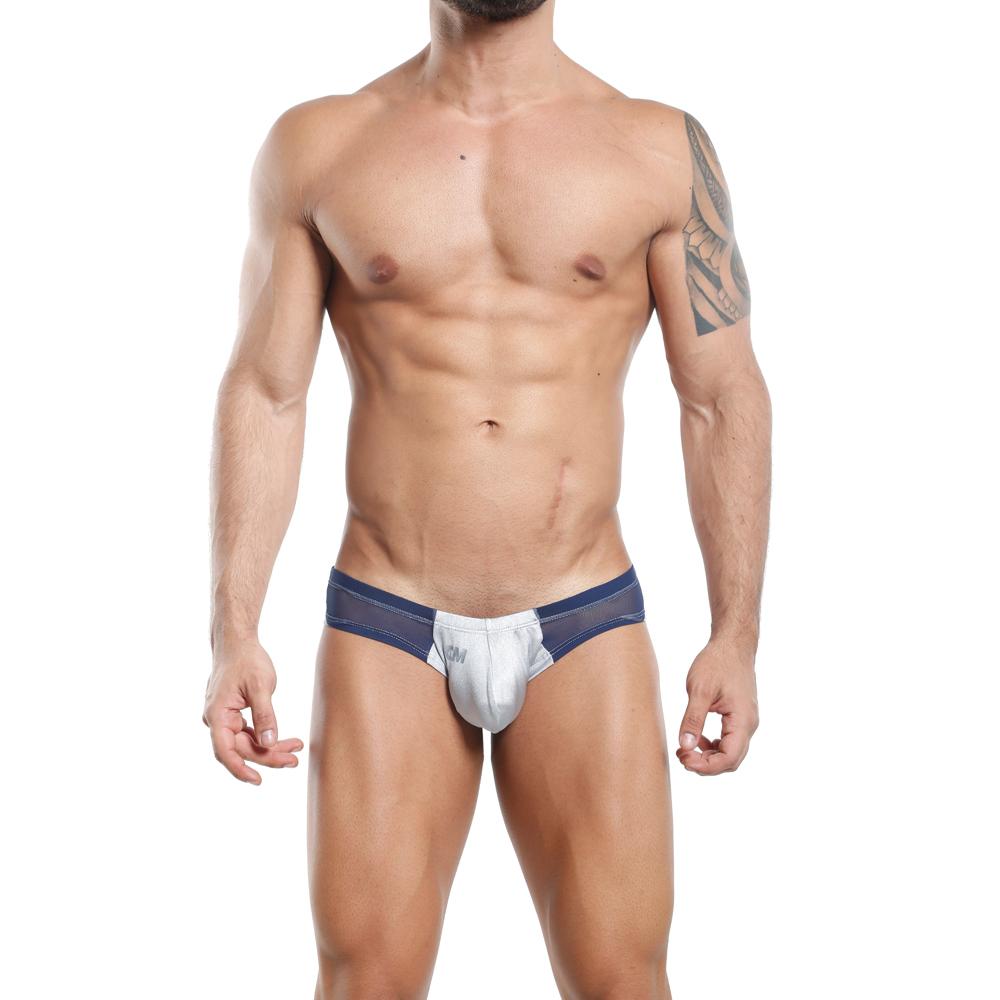 A person wearing the Cover Male Slip Thong in blue and gray stands with hands by their sides, displaying a tattoo on their right arm. The stylish design and fabric fusion provides supportive hold. The setting is a plain white backdrop.