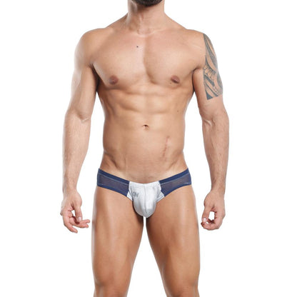 A person wearing the Cover Male Slip Thong in blue and gray stands with hands by their sides, displaying a tattoo on their right arm. The stylish design and fabric fusion provides supportive hold. The setting is a plain white backdrop.