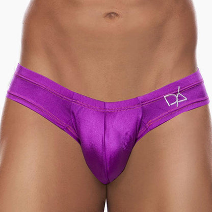 A person stands against a plain background wearing the Daniel Alexander Emotion Slip Thong in bright purple with a cheeky back design and a small logo. Made from a premium fabric blend, the thong has a shiny, satin-like texture. Only the midsection is visible.