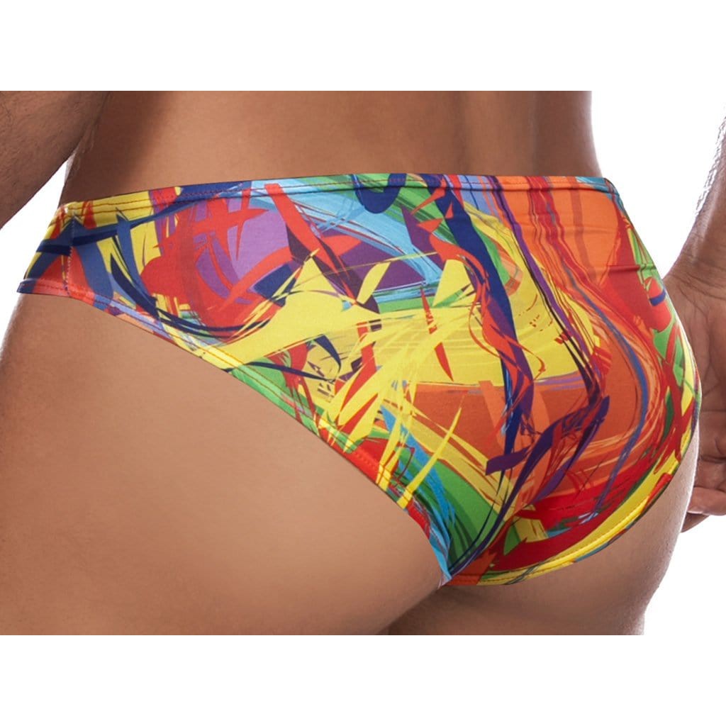 A person wearing vibrant swim trunks with a multicolored pattern showcasing red, yellow, blue, and green strokes. The Daniel Alexander Explosion Slip Bikini features modern side prints and a contouring pouch for enhanced design.