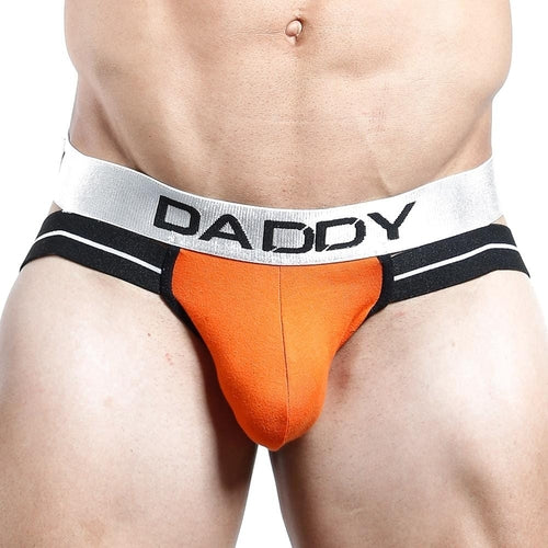 A close-up of a muscular man wearing the Daddy brand “Daddy Jockstrap – Wear the Title, Own the Throne” featuring orange and black design, V-shaped pouch, and a wide white waistband boldly printed with DADDY.