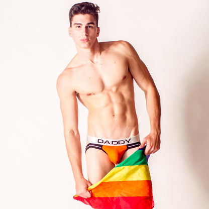A young man poses against a plain background, holding a rainbow pride flag and wearing the Daddy Jockstrap by Daddy—featuring a bold waistband so you can wear the title and own the throne.