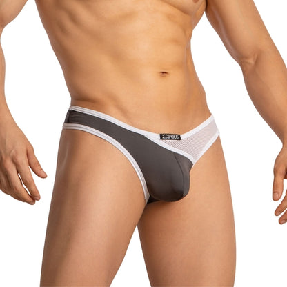 A person poses with hands at their sides, wearing Edipous Mens Thong with an asymmetrical cut in two-tone gray and white. The stylish design features mesh fabric on the sides and a branded logo on the waistband.