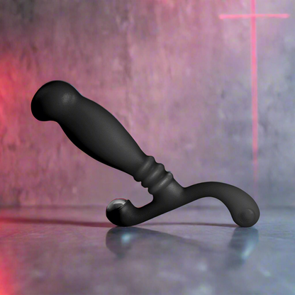 A Nexus Glide Prostate Massager by Nexus, featuring a black ergonomic S-shaped design with a curved handle, rests on a reflective surface. The background displays a textured wall with a subtle red cross projection, highlighting its perineum stimulation allure.
