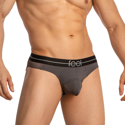 Sporting a fit physique, he models the Feel Thongs for Guys by Feel, featuring grey briefs with a black waistband. Mesh panels on the sides add style and comfort, highlighting the perfect blend of functionality and flair in mens thongs.