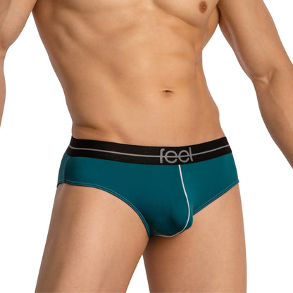 A person wearing Feels teal thongs for men with a black waistband featuring the brand name. The image highlights their torso and upper thighs, showcasing the minimal coverage design made from a luxurious fabric blend.
