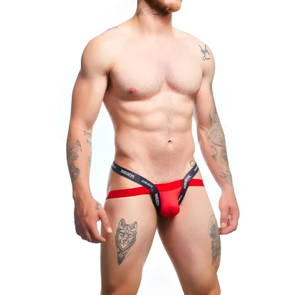 A muscular, tattooed man models the Good Devil Sexy Men Jockstrap in bright red with a black waistband. The premium fabric blend highlights his physique against a simple white background, enhancing the artwork of his arm and leg tattoos.