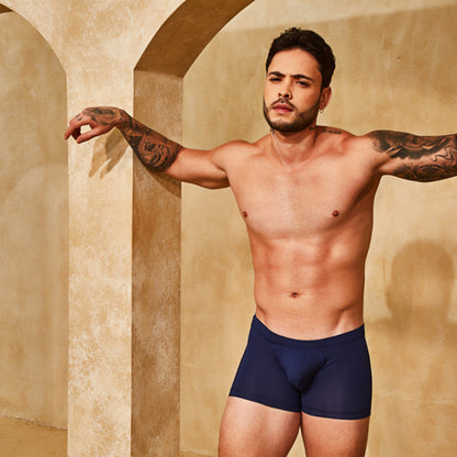 A tattooed man poses shirtless against a beige wall with archways, wearing navy Intymen Good Trunks that fit perfectly. His arms are outstretched as he gazes sideways, embodying the premium fabrics of Intymen.