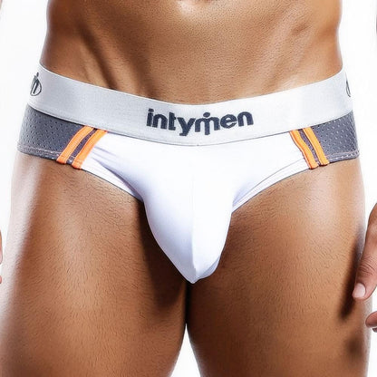 A man models the Intymen Vibrant Attitude Bikini Brief in white and gray with orange trim. Designed for comfort, they feature a contoured center pouch for enhanced fit and support. Intymen is displayed on the waistband, with a white backdrop highlighting the lower torso.