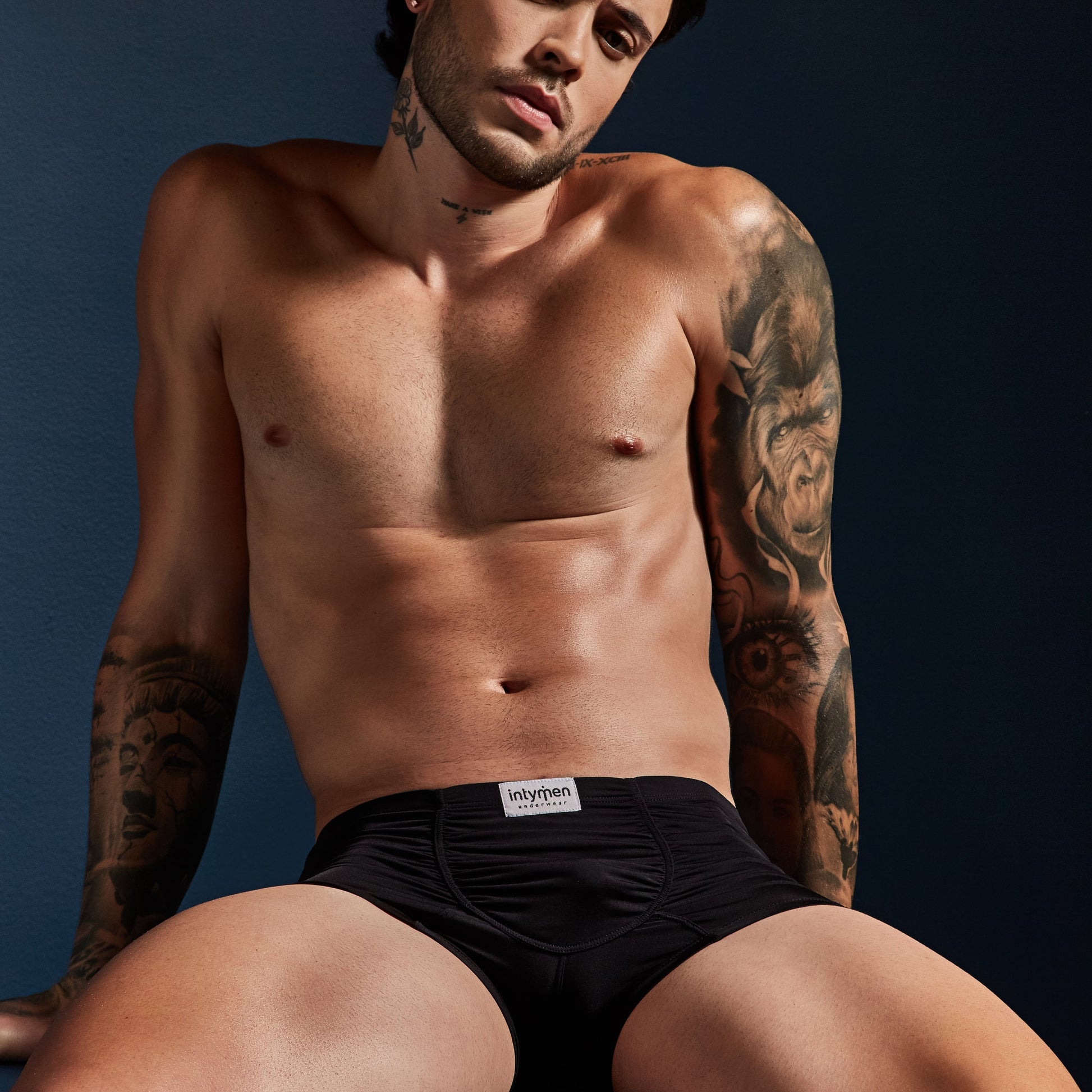 A shirtless individual against a dark background showcases the Intymen Star Pouch Boxer Trunk Underwear in black, featuring a spacious pouch and soft, stretchy fabric for comfort. The Intymen brand is visible on the waistband, with tattoos on their arms and torso.