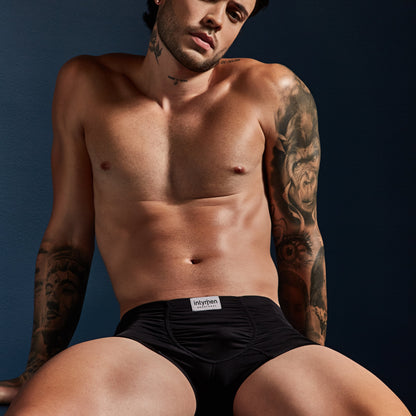 A shirtless individual against a dark background showcases the Intymen Star Pouch Boxer Trunk Underwear in black, featuring a spacious pouch and soft, stretchy fabric for comfort. The Intymen brand is visible on the waistband, with tattoos on their arms and torso.