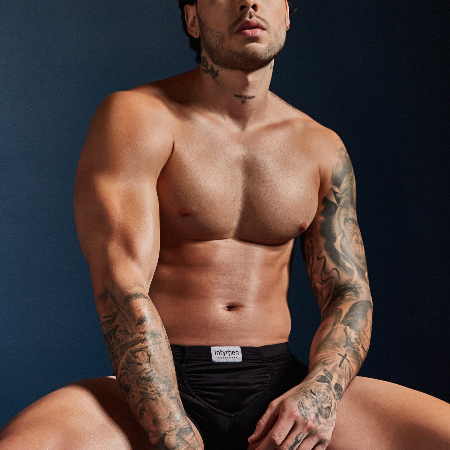 A tattooed person sits shirtless on a dark surface, wearing Intymen Star Pouch Boxer Trunk Underwear made of soft, stretchy fabric, set against a dark blue background.