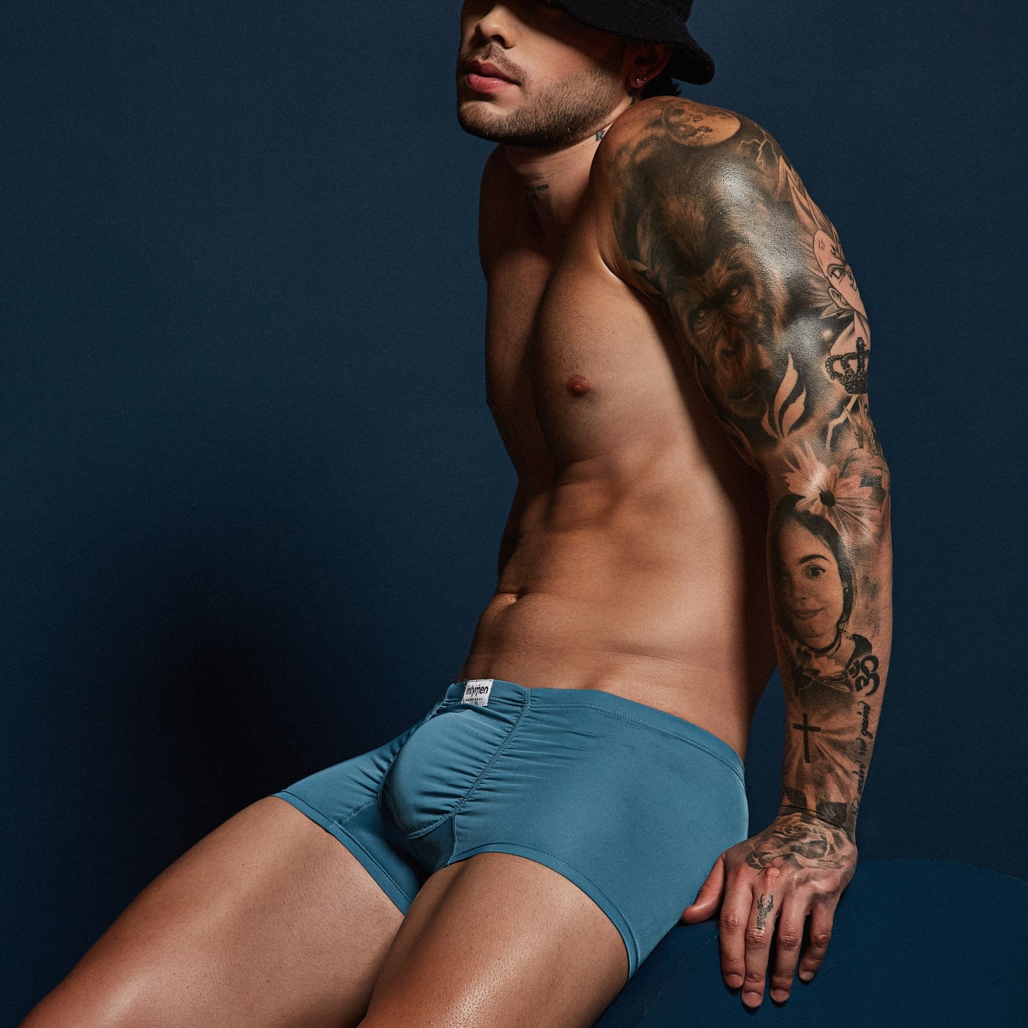 A shirtless man in Intymen Star Pouch Boxer Trunk Underwear, with a spacious pouch and soft, stretchy fabric, poses against a dark background. He wears a black hat and flaunts his detailed tattoo sleeve on his right arm.