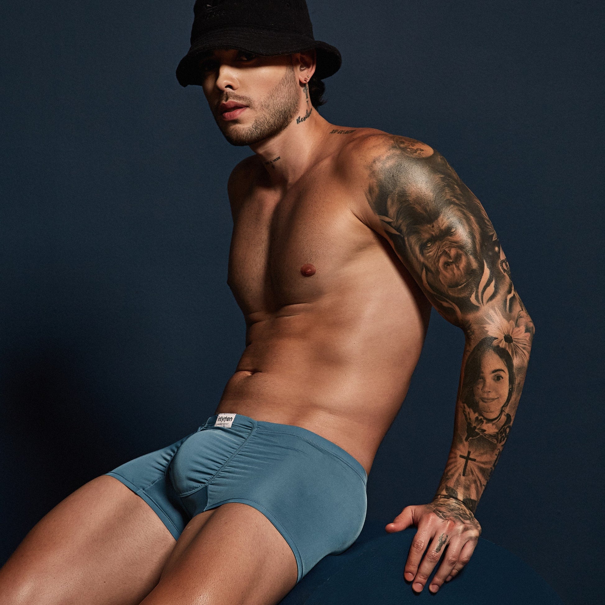 With tattooed arms, a man poses against a dark background, wearing a black bucket hat and Intymen Star Pouch Boxer Trunk Underwear in teal. Made from soft, stretchy fabric, he sits with one knee up, gazing thoughtfully to the side and showcasing the spacious pouch perfectly.