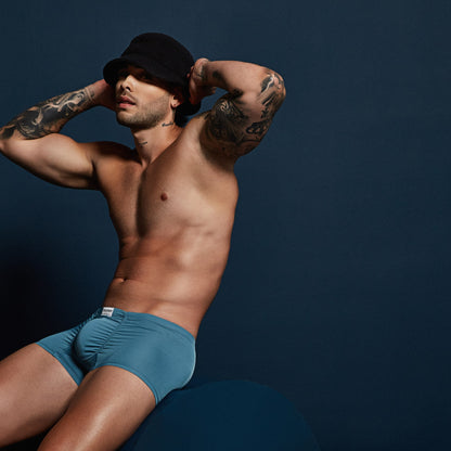 A shirtless, tattooed man wears Intymen Star Pouch Boxer Trunk Underwear made of soft, stretchy fabric and a black bucket hat. Seated against a dark blue background, he gazes aside with hands on the hat. The boxer offers a spacious pouch for comfort and style.