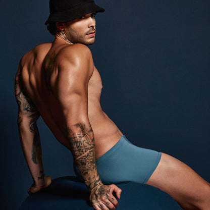 A tattooed man in a black hat and Intymen Star Pouch Boxer Trunk Underwear sits on a round blue object against a dark background, turned slightly to the side to reveal his muscular arms and back.
