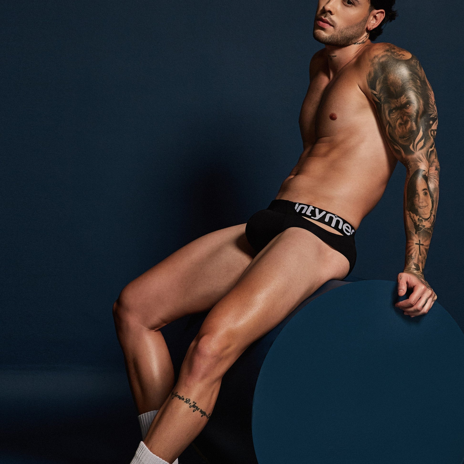 A tattooed, shirtless individual sits on a large, dark cylinder wearing Intymens True Brief Underwear, crafted for comfort with a premium fabric blend and slim fit. Theyre also wearing white socks against a dark backdrop.