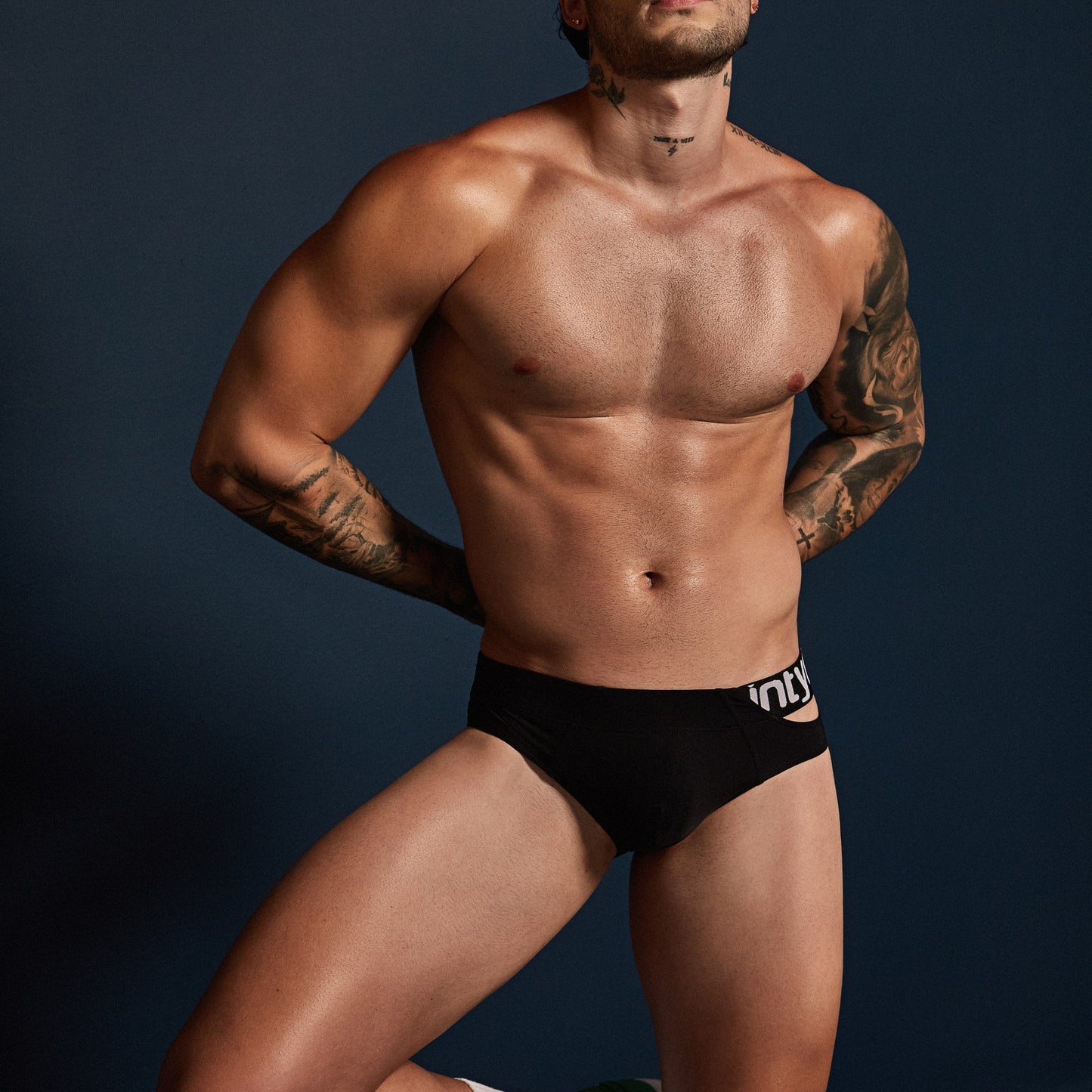 A shirtless man models Intymen True Brief Underwear against a dark backdrop, with the slim fit accentuating his arm and neck tattoos; his hands rest behind his back.