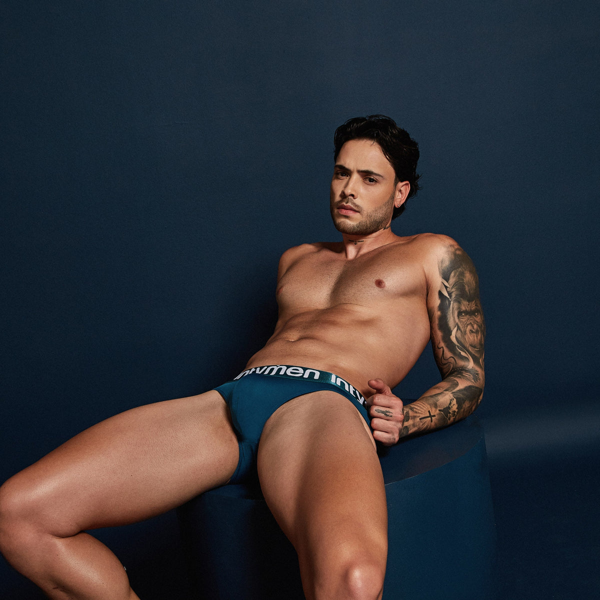 Man seated against a navy backdrop wearing the Intymen Sky Jockstrap in Blue Petroleum, intymen logo in white on the waistband.