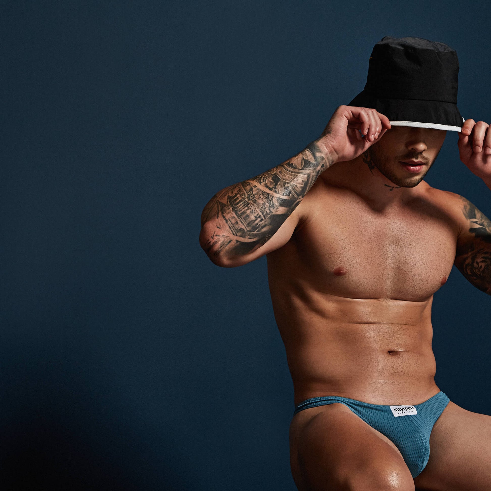 A tattooed, shirtless man in Intymen Basic Mesh Thong INK017 and a black bucket hat adjusts its brim while seated against a dark blue background.